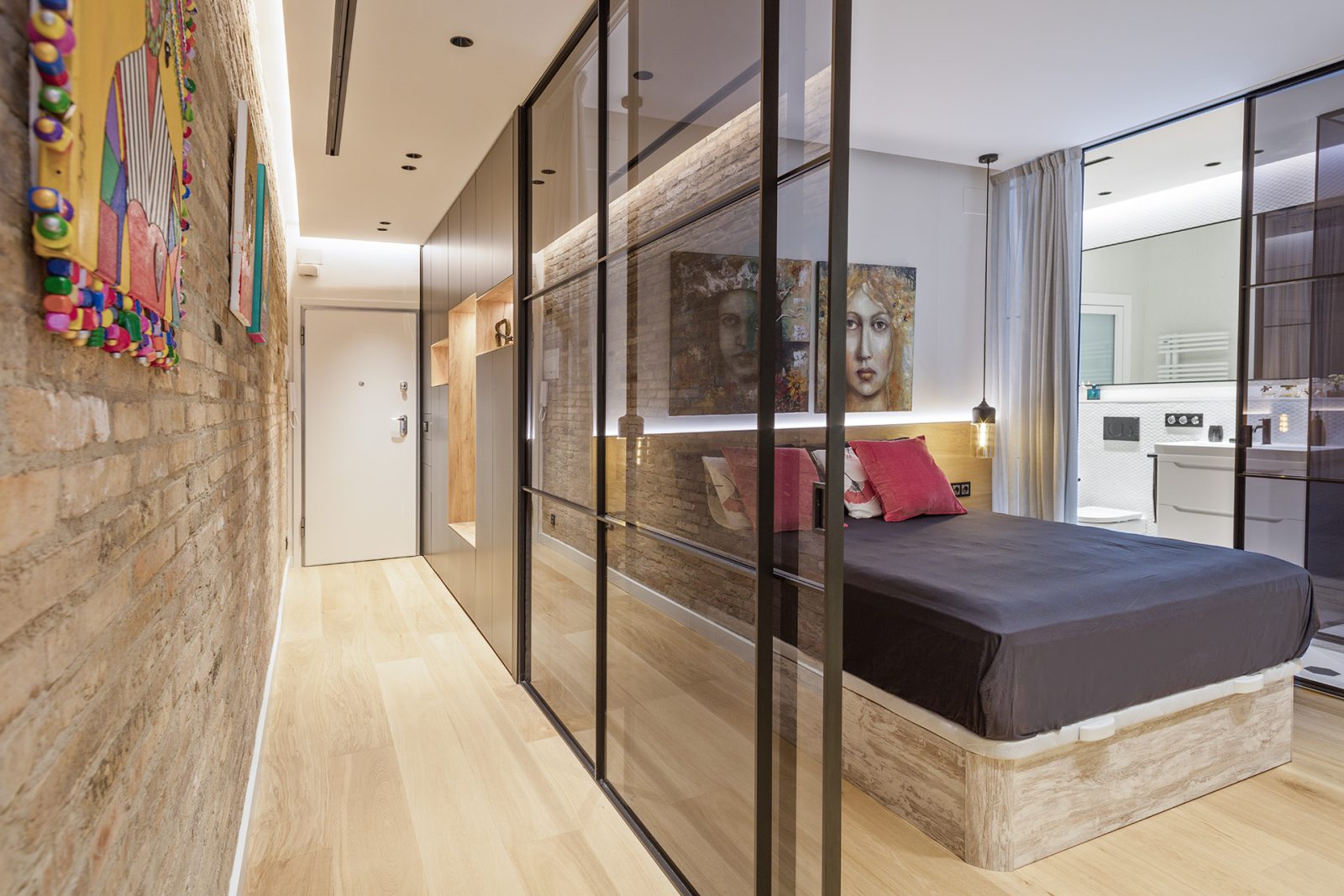 Dormitorio con cerramiento acristalado, perfilería negra, ladrillo visto e iluminación integrada en un apartamento de Sitges.
